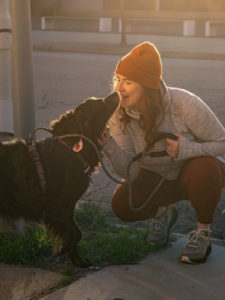 Amanda kneeling on a sidewalk while kissing a black retriever