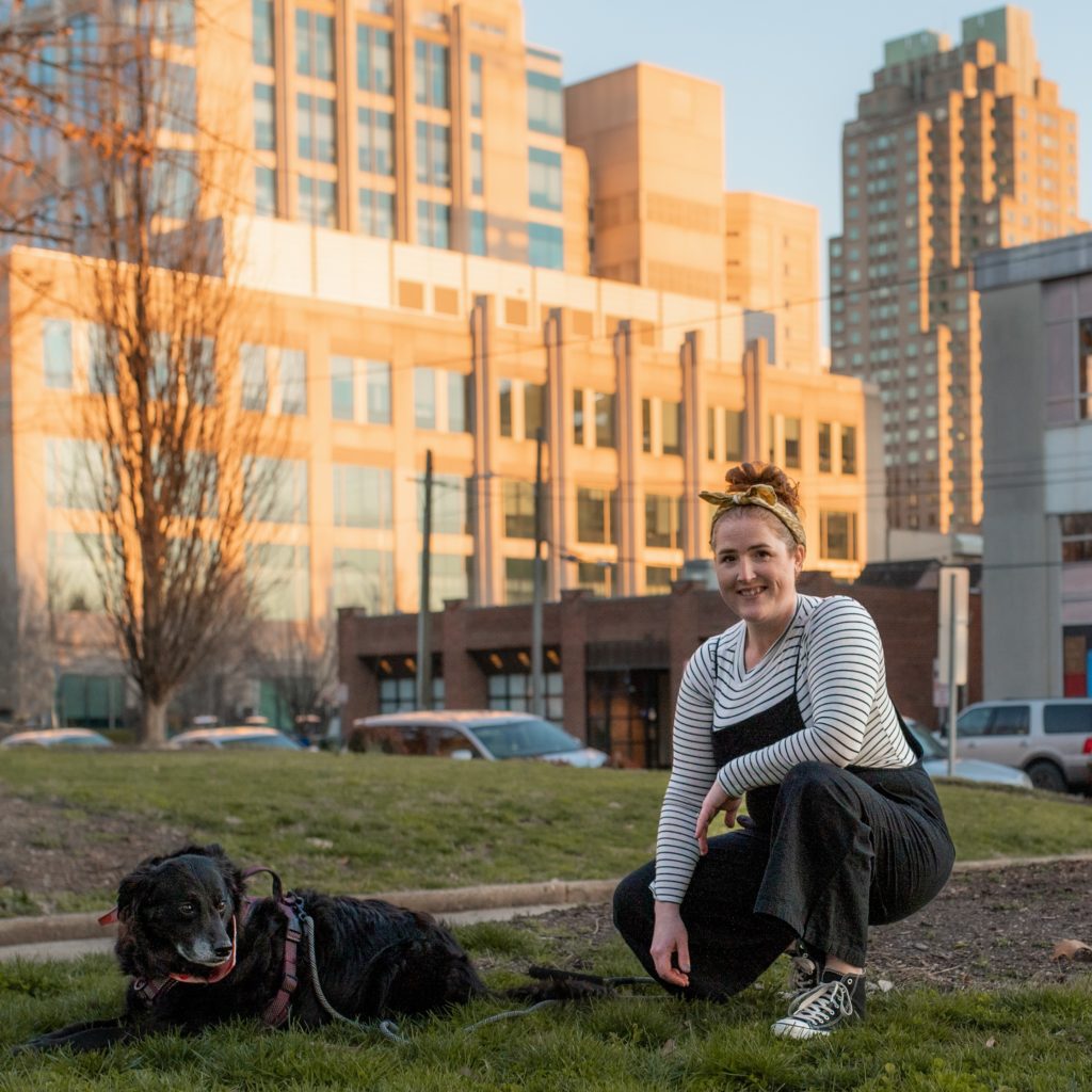 Amanda kneeling in a downtown park with a black retriever next to her and Raleigh buildings in the background