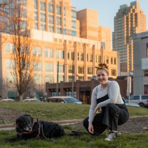 Amanda kneeling in a downtown park with a black retriever next to her and Raleigh buildings in the background
